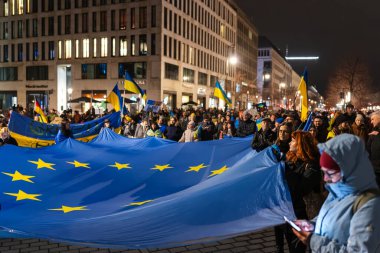 Berlin, Almanya 24 Şubat 2026: Büyük bir kalabalık, Ukrayna yanlısı bir gösteride şehir merkezi meydanına uzanan büyük bir Avrupa Birliği bayrağı taşıyor. Toplantının üzerine sayısız Ukrayna mavi ve sarı bayrakları asıldı.