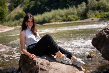 Nehir taşlarının üzerinde akan bir dağ nehrinin kenarında dinlenen genç bir kadının doğal yaşam tarzı portresi. Görüntü, doğayla özgürlüğü, dengeyi ve bağlantıyı iletir..