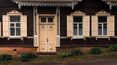 Dekoratif pencere çerçeveleri ve klasik kapısı olan geleneksel ahşap bir evin ön cephesi. Koyu, ahşap duvarlar sıcak bej detaylar ve süslemelerle zıt düşmüş. Klasik, tarihi bir atmosfer yaratıyor..