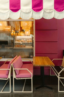 Pembe kafe terası, rahat bir açık hava koltuğu ve sıcak iç ışıkları var. Şık sandalyeler ve ahşap masa kahve molası için davetkar bir ortam yaratır..