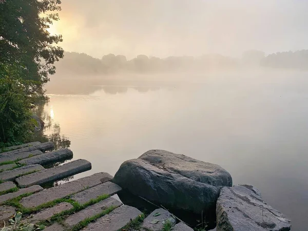 Sonbaharda Şafakta Sakin Göl 'ün Kıyısında Güneş Işığı Sis ve Kayalıklar. Sisli bir sabahta huzurlu, atmosferik bir manzara. Soldaki alçak güneş, durgun göl suyunun üzerinde süzülen yoğun siste altın bir parıltı saçar.. 
