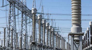 Açık mavi gökyüzü altında çok sayıda yüksek voltajlı yalıtkan, elektrik hatları ve iletim kulelerinin yer aldığı bir elektrik santralinin geniş bir görüntüsü.