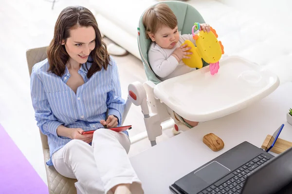 smiling brunette woman with graphic tablet in hands sitting at laptop, child with toy sitting next to her, Working from home, remote work at home with children