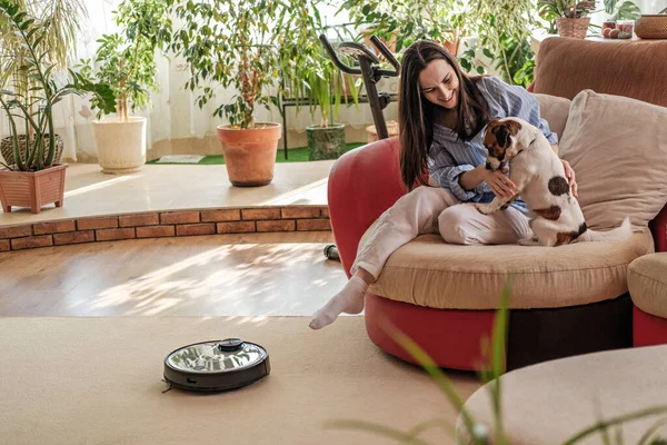 Robotik süpürge halıda, mavi gömlekli kadın köpekle oynuyor, Jack Russell Terrier kanepede doğuruyor, yaşam konseptinin tadını çıkarıyor, rahat ve rahat ev, akıllı cihaz.