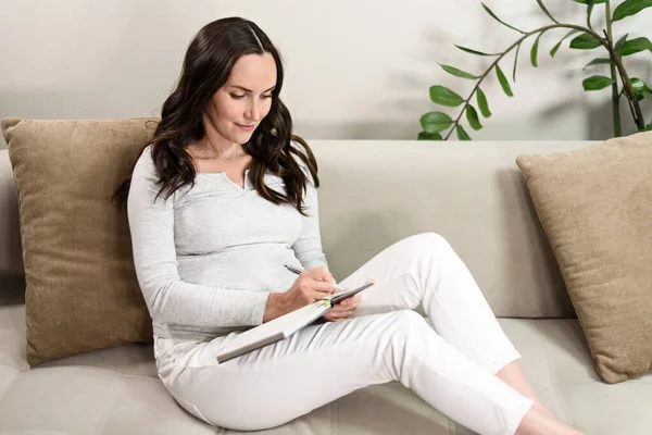 Έγκυος Γυναίκα Γράφει Στο Σημειωματάριο Γκρι Μπλούζα Κάθεται Στον Καναπέ — Φωτογραφία Αρχείου