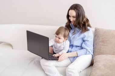 Bebekli beyaz bir kadın dizüstü bilgisayarda çizgi film seyrediyor. Kanepede oturuyor. Evde çocuklarla uzaktan çalışıyor.