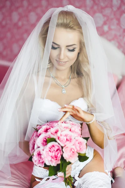 Schöne sexy junge Braut Unterwäsche sitzt auf einem Bett mit einem Blumenstrauß in den Händen und mit einem Schleier auf dem Kopf. Morgen die Braut. — Stockfoto