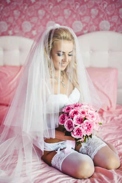 Schöne sexy junge Braut Unterwäsche sitzt auf einem Bett mit einem Blumenstrauß in den Händen und mit einem Schleier auf dem Kopf. Morgen die Braut. — Stockfoto