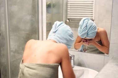 Kafasında havlu olan genç bir çocuk banyo lavabosunda yüzünü yıkıyor.