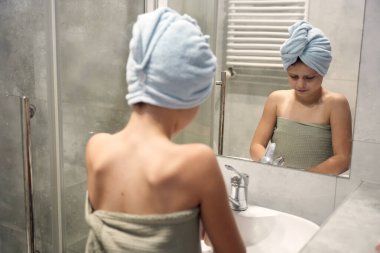 Kafasında havluyla bornoza sarılmış çocuk lavaboda dikilip yansımaya bakıyor.