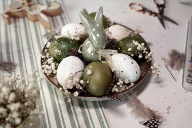 Hassas çiçekler ve tüylerle süslenmiş benekli beyaz ve yeşil yumurtalarla dolu bir tabak, merkezdeki şirin bir tavşana odaklanmış..