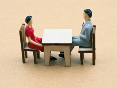 İki küçük figür, kırmızılı bir kadın ve gri elbiseli bir adam, basit bir masada karşılıklı oturuyorlar. Bir sohbete katılmış gibi görünüyorlar.