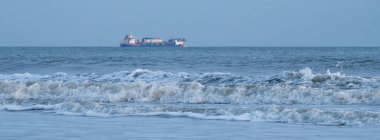 Alacakaranlıkta kumlu bir sahile vuran hafif dalgalar gibi uzakta bir kargo gemisi görülüyor. Sakin atmosfer huzurlu bir sahil manzarası yaratır.