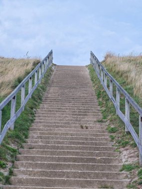 Çerçevelenmiş yeşil çimenler ve ahşap korkuluklar ile bir zirveye çıkan uzun bir beton merdiven seti. Başımızda, bulutlarla dolu engin bir gökyüzü dingin atmosfere katkı sağlar.