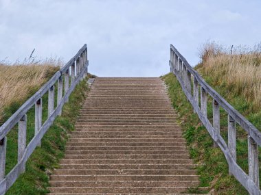 Güçlü taş basamaklar, doğal yeşilliklerle çevrili çimenli bir yamaçtan yükselir. Bir ahşap korkuluk her iki tarafta bulutlu bir gökyüzünün arka planına doğru uzanır.