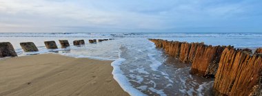 Weathered wooden pilings emerge from the sandy beach as gentle waves roll in. The calm ocean reflects the soft colors of the sky at sunset, creating a peaceful atmosphere