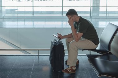Havaalanı kapısında bavuluyla oturan genç bir adam uçuşu bekliyor, akıllı telefonuna bakıyor. Yolcu uçuş gecikmesinden dolayı üzgün. Modern hava yolculuğu konsepti. Yüksek kalite fotoğraf