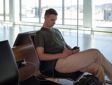 Havaalanı kapısında telefonu şarj eden genç bir adam uçuş beklerken akıllı telefon kullanıyor. Modern terminalde rahat seyahat konsepti. Yüksek kalite fotoğraf
