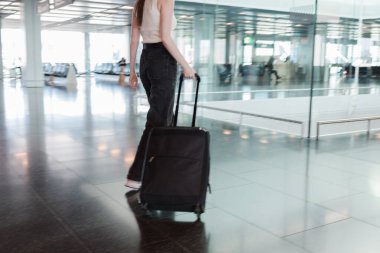 Havaalanı terminalinde siyah bavullu hareket bulanıklığıyla yürüyen genç bir kadın. Bağımsız seyahat kavramı, modern yaşam tarzı ve kusursuz hareket kabiliyeti. Hareket halindeki fotoğraf, yumuşak odaklanma. Yüksek kalite fotoğraf