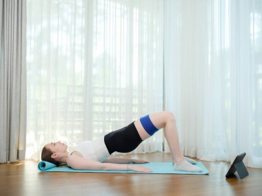 Evde yoga minderinde direniş bandıyla egzersiz yapan genç bir kadın. Spor anlayışı, sağlıklı yaşam tarzı ve online egzersiz. Tablet üzerinde sanal eğitimi takip ederken kadın kalça köprüsü yapıyor