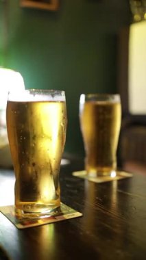 İki bardak taze dökülmüş bira bir bar masasında dikilirken arka plandan ön plana sorunsuzca kayıyor, kabarcıklar ve davet edici bira dokusu, 1080p 60 fps