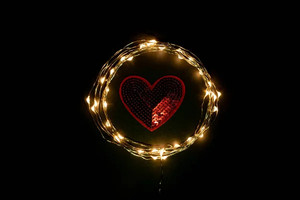 Red Heart in the circle of holiday celebration light garland on black background. Romantic Valentines day cards, invitations or posters. Dark mood