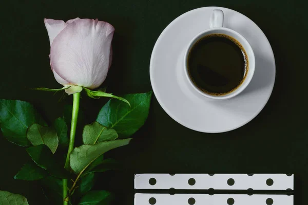 Cup of black coffee, notebook and pink rose on black background, Top view, copy space. Toned