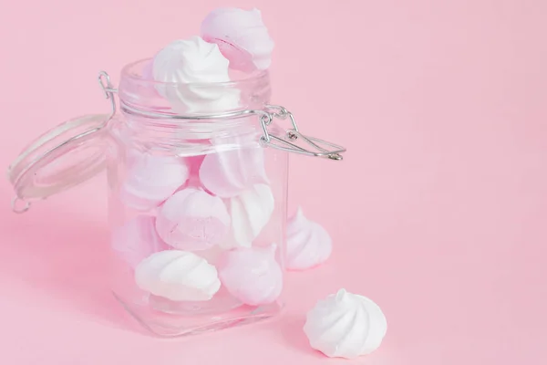 White and pink twisted meringues in a glass jar on pink background. French dessert prepared from whipped with sugar and baked egg whites. Greeting card with copy space