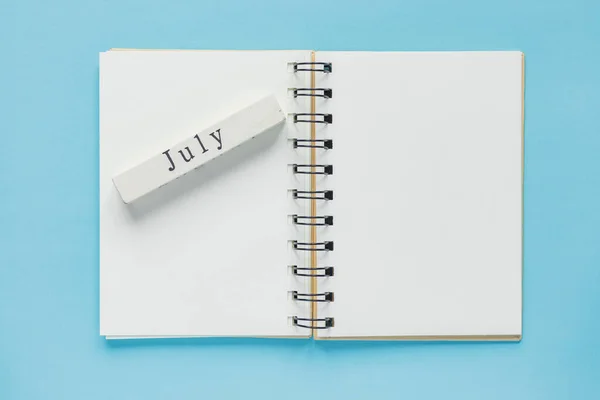 Clean spiral note book for notes and messages and july wooden calendar bar on blue background. Minimal business flat lay