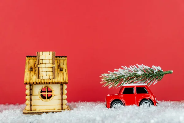 Small red toy car carrying spruse tree on a snowe road next to wooden hous. Festive christmas greeting card