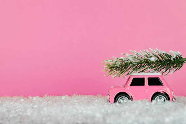 Small pink toy car carrying spruse tree on a snowy road. Festive christmas greeting card