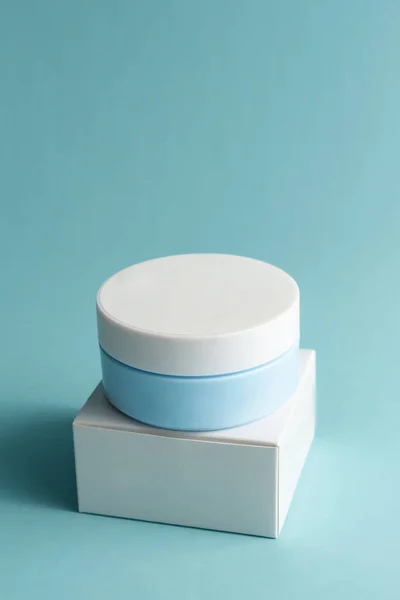 Face cream jar with plastic cap and whire carton box, package on blue background. Mockup unbranded cosmetology product.