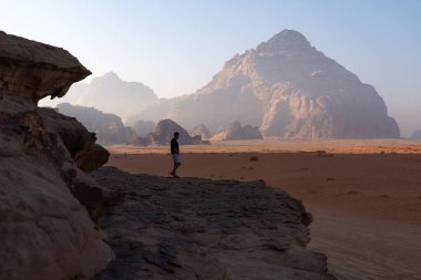 Çöle bakan kayalık bir çıkıntıda yürüyen bir erkek turistin yan görüntüsü. Ürdün 'deki Wadi Rum' da sabah ışığında muazzam kumtaşı dağları yükselir..