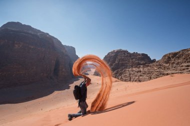Erkek turist bir kum tepesinde diz çöküp havaya kırmızı kum fırlatarak dinamik bir kavis oluşturuyor. Güneşli bir günde, Ürdün 'ün Wadi Rum dağlarının arasında eğlenceli bir an..
