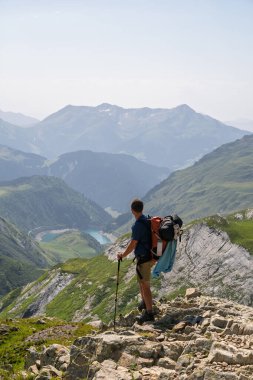 Kayalık bir tepenin üzerinde duran bir erkek yürüyüşçünün arka görüntüsü aşağıda bir su deposu olan geniş bir dağ vadisine bakıyor. Mont Blanc turu sırasında Fransız Alplerinde çekilmiş, manzaranın boyutunu vurguluyor..