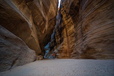 Siq Kanyonu 'nun devasa kumtaşı duvarlarının altında duran erkek turistin geniş açılı görüntüsü. Dar geçit, Ürdün 'ün Petra antik şehrine çıkıyor. Dramatik ışıklandırma ölçeği vurguluyor.