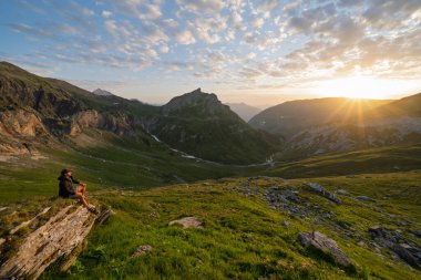 Kayalık bir çıkıntıda oturan bir erkek yürüyüşçünün, La Gittaz yakınlarındaki engin yeşil Alp Vadisi 'nde gün batımını seyrederken yan görüntüsü. Mont Blanc turu sırasında Fransız Alplerinde dramatik gölgelerle çekildi..