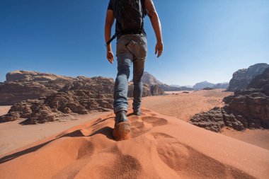 Kırmızı kum tepeciğinin tepesinde yürüyen erkek bir turistin alçak açılı arka görüntüsü. Wadi Rum, Ürdün 'deki çöl dağlarına karşı kuma batan botlara odaklan. Parlak güneşli bir gün.