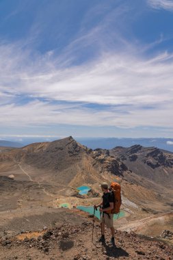 Tepenin tepesinde duran bir erkek yürüyüşçünün canlı turkuaz Zümrüt Gölleri 'ne bakışının yan görüntüsü. Yeni Zelanda 'daki Tongariro Alp Dağları' nda çekildi. İnce bulutlu mavi gökyüzünün altında volkanik bir manzara.