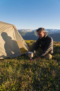 Çadırının dışındaki ocakta yemek pişiren ceketli bir erkek yürüyüşçü. Gölgesi çadır kumaşına yansıyor. Norveç 'in Hardangerfjord tepesindeki dağlarda vahşi bir kamp. Altın saat güneş ışığı.
