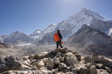 Turuncu bir sırt çantasıyla bir kaya yığınının üzerinde duran erkek bir yürüyüşçünün, Nuptse Dağı 'nın devasa karlı zirvesine baktığının arka görüntüsü. Sagarmatha Ulusal Parkı, Nepal 'deki Everest Ana Kamp Yürüyüşü' nde çekildi..
