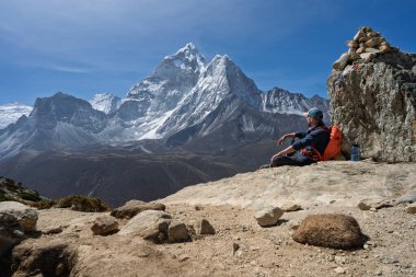Ama Dablam 'ın ikonik keskin zirvesine hayran bir erkek yürüyüşçünün taş bir cairn' in yanında oturuşu. Nepal Himalayaları 'ndaki Everest Ana Kampı' nda Dingboche 'un yukarısındaki bir sırtta çekildi. Güneşli bir gün.