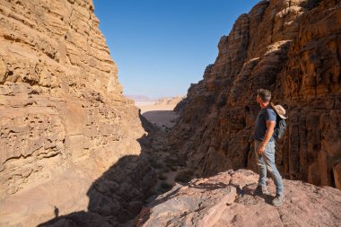Kayalık bir çıkıntıda duran bir erkek turistin derin, dar bir kumtaşı kanyonuna bakışını. Vadi, Ürdün, Wadi Rum 'daki çöl ovasına açılıyor. Sert jeolojik oluşumlar.