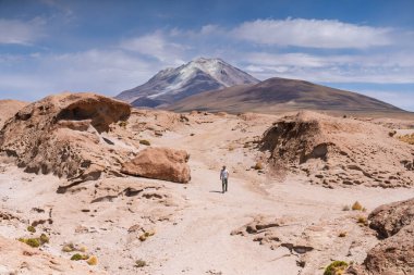 Antik kaya oluşumlarının uçsuz bucaksız, çorak bir arazisinde yürüyen bir yürüyüşçünün geniş açılı görüntüsü. Volkan Uturuncu 'nun yüksek irtifada Bolivya Altiplanosu' nda görünmesi muhtemel büyük bir stratovolcano..
