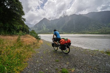 Yeni Zelanda 'daki Haast Nehri' nin yanındaki çakıllı yolda bisiklet süren bir erkek bisikletçinin arka görüntüsü. Güney Adası 'ndaki sisli dağlara karşı pannierlerle bisiklet sürüyor.
