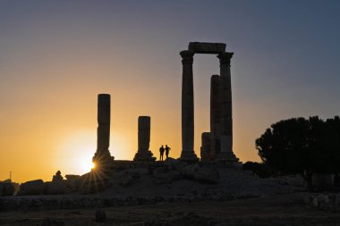 Herkül Tapınağı 'nın devasa Roma sütunları arasında duran iki siluet. Ürdün 'deki Amman Kalesi Jabal al-Qala' da altın bir günbatımı gökyüzü tarafından aydınlatıldı. Dramatik tarihi sahne.