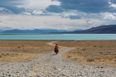 Kurak Patagonya bozkırlarında uzak bir çakıllı yolda dolu bir bisiklete binen bir bisikletlinin arka görüntüsü. Canlı turkuaz Lago Argentino uçsuz bucaksız, bulutlu gökyüzü Arjantin 'in altında ufukta hüküm sürüyor..