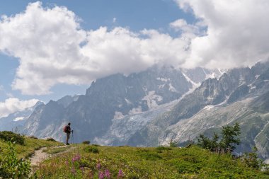Çiçekli bir patikada duran bir erkek yürüyüşçünün, Mont Blanc 'ın devasa granit tepelerine ve buzullarına bakışının yan görüntüsü. Tour du Mont Blanc yürüyüşü sırasında İtalyan Alpleri 'nde çekilmiş Val Ferret. Zirvelerdeki bulutlar.