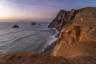 Paracas Ulusal Rezervi 'nin engebeli, kurak uçurumları ve deniz yığınları üzerinde dramatik bir gün batımı, Peru. Yumuşak pastel ışık, Pasifik Okyanusu 'yla buluşan çöl kıyılarını aydınlatır..