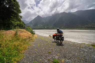 Yeni Zelanda 'nın Haast Nehri' nin yanındaki gevşek çakıl yolu üzerinde bisiklet süren bir erkek bisikletçinin ön görüntüsü. Teçhizatını Güney Adası 'ndaki sisli dağlarla karşılaştırmak için duruyor.